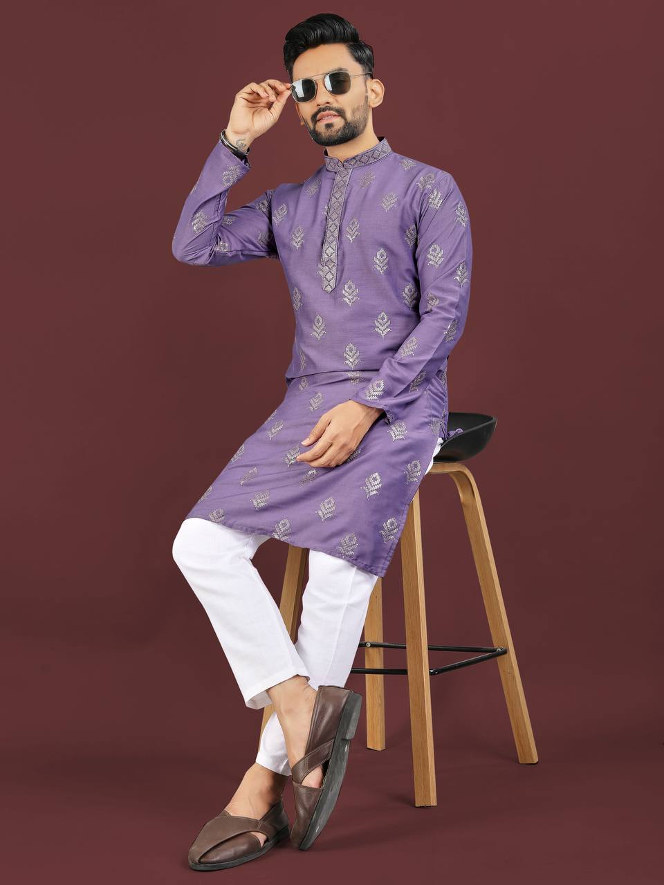 Man wearing a lavender jacquard kurta set with white pants and brown shoes sitting on a stool