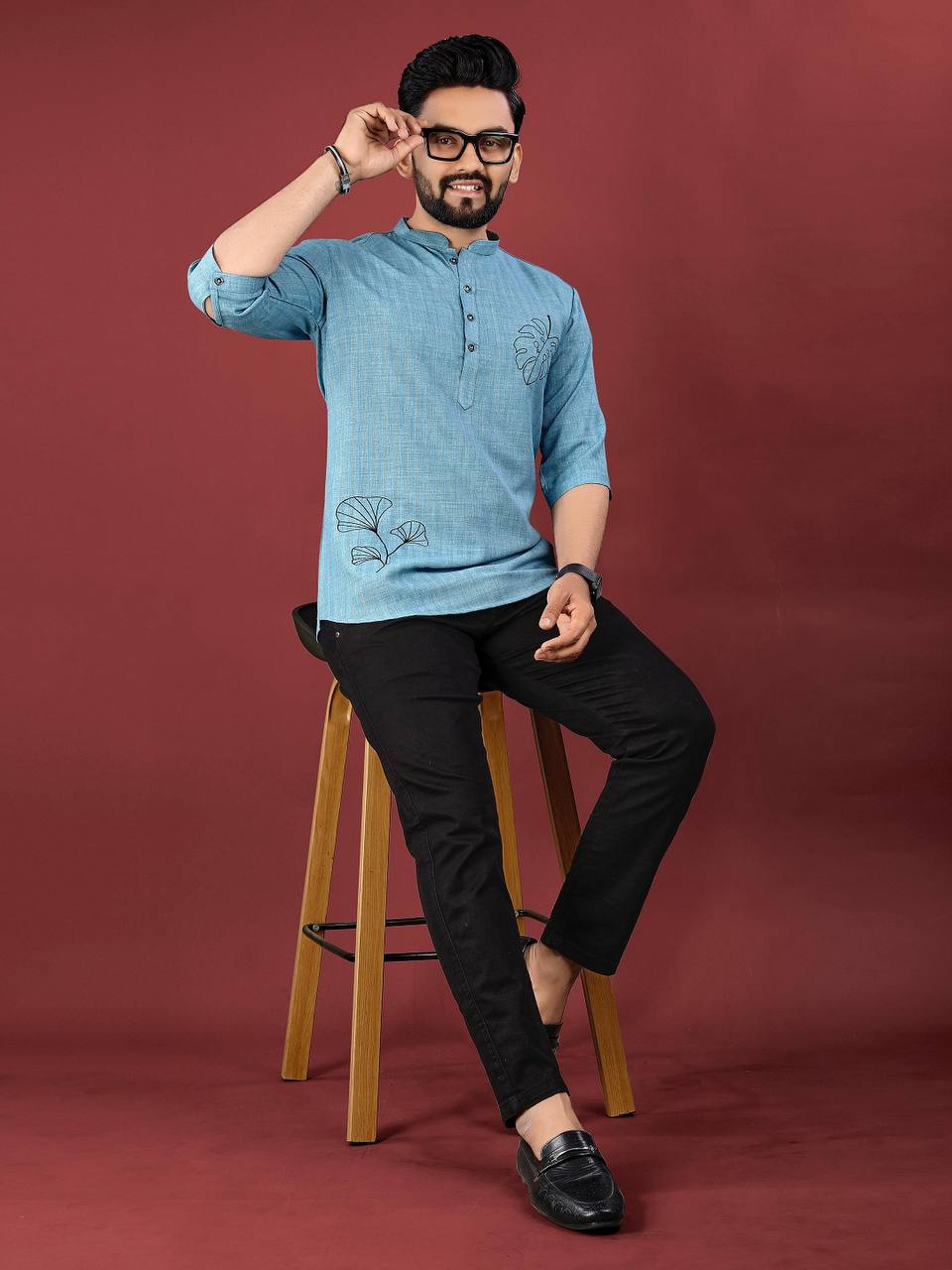 Man wearing a blue ocean leaf embroidered kurta sitting on a wooden stool against a maroon background