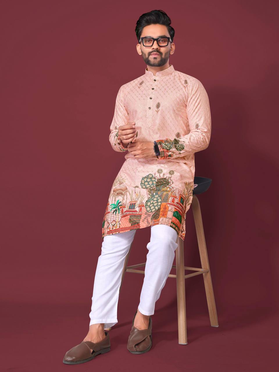 Man wearing peach print kurta for men with white pants and brown shoes sitting on stool